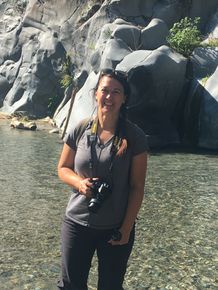 Tawnya Bujold smiling and holding a camera in her right hand, standing in front of water and boulders