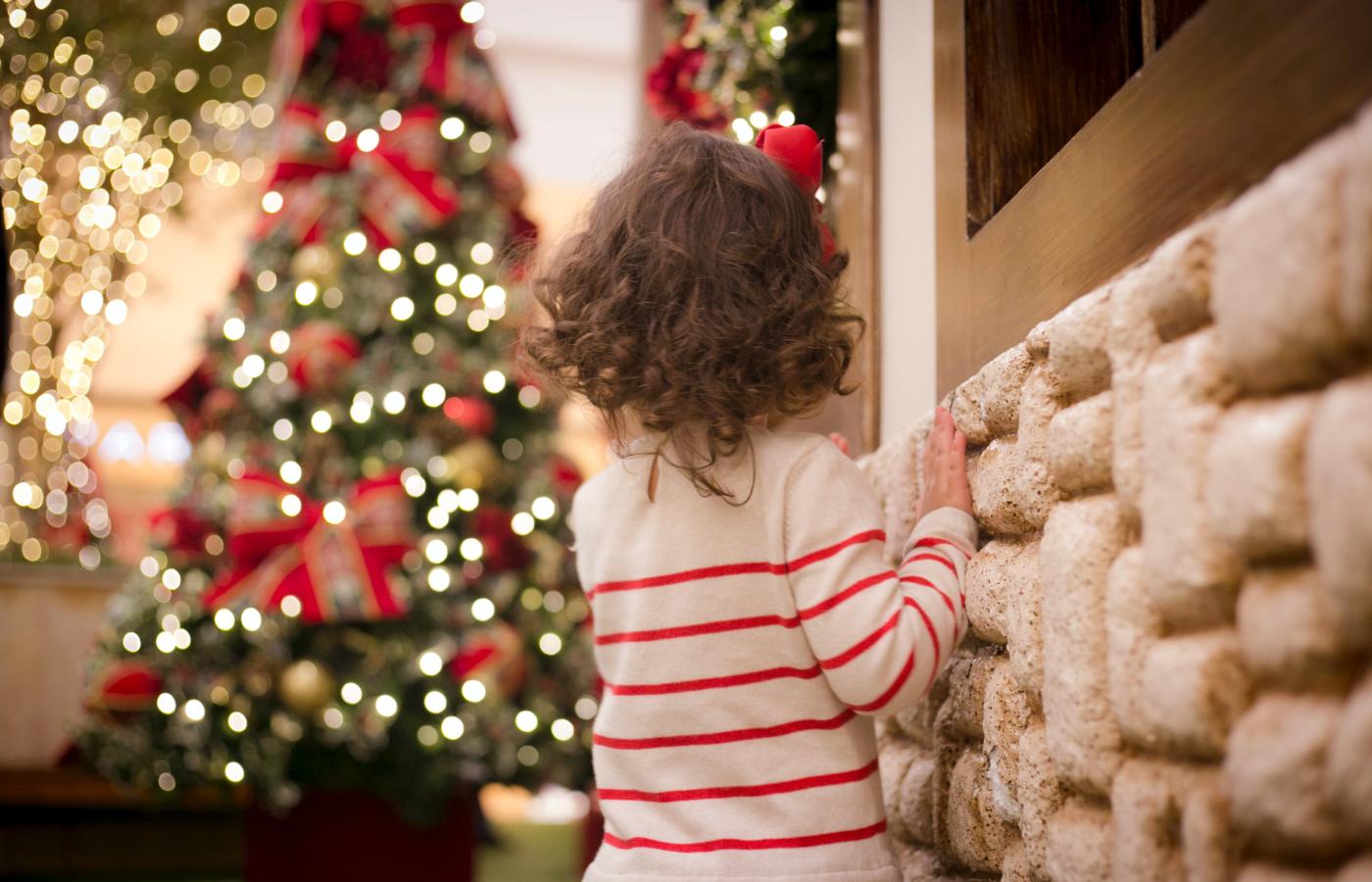 Small child standing next to a white brick wall looking out into the distance of a lit christmas tree adorned with white lights and red ribbons